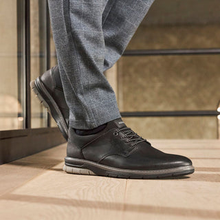 Black leather shoes worn with gray pants on a tiled floor.